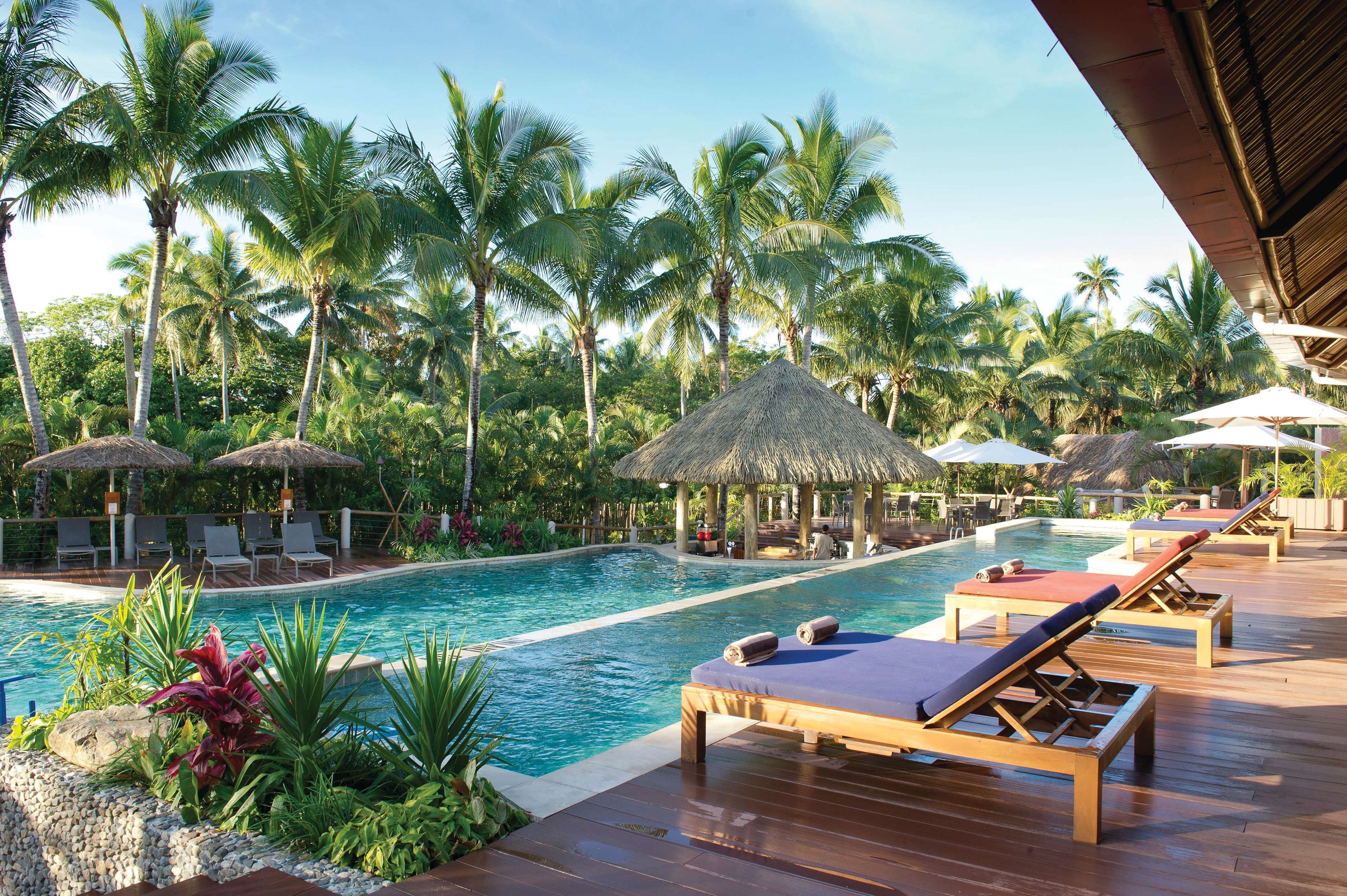A pool area with lounge chairs, palm trees, and a thatched-roof structure.