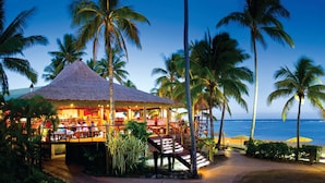 Un restaurante frente a la playa con techo de paja, palmeras y vista al océano.