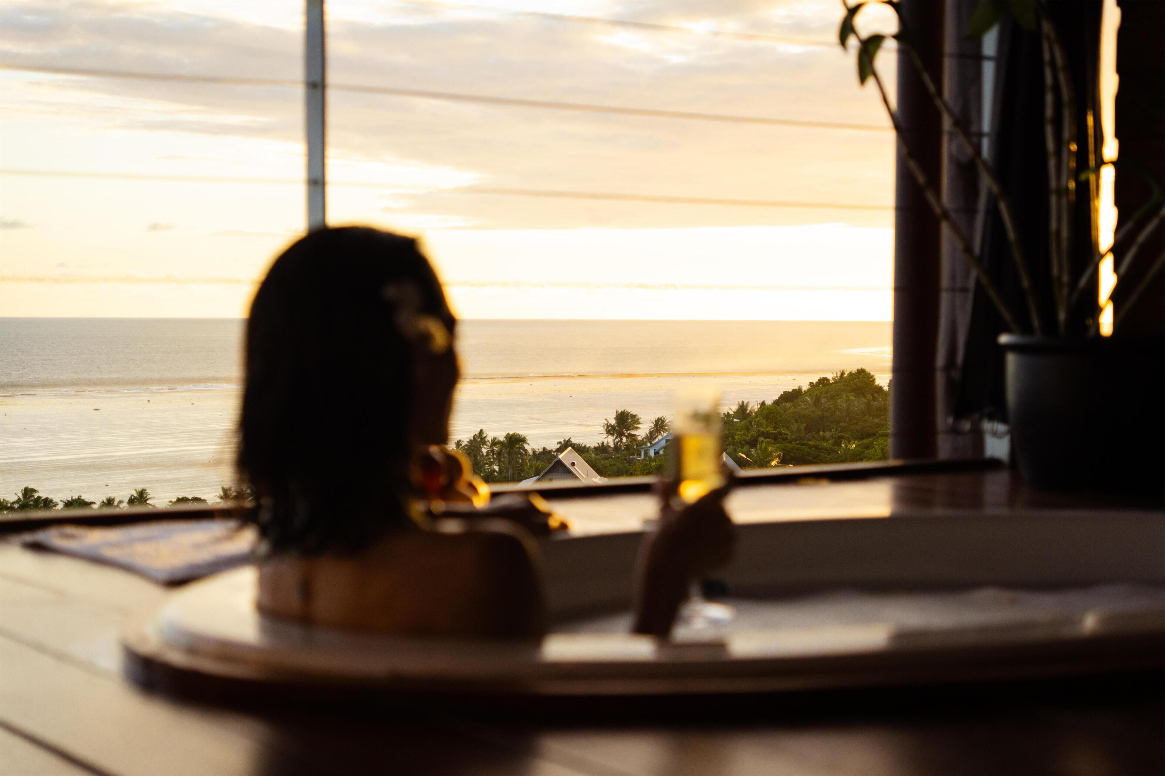 Une personne est assise près d’une fenêtre donnant sur une étendue d’eau, avec une plante et une table au premier plan.