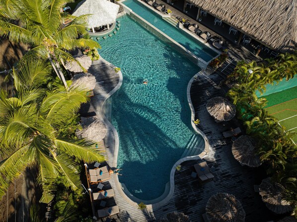 Vista aérea de una piscina en un resort, con palmeras, sillas de descanso y una estructura de techo de paja.