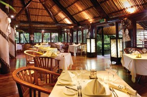 Un restaurante con varias mesas dispuestas para comer, sillas de madera y un techo de paja.