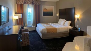 A hotel room with a large bed, a desk with a lamp, a chair, a painting on the wall, and a window with curtains. - Days Inn by Wyndham Mesquite Rodeo TX (Mesquite)