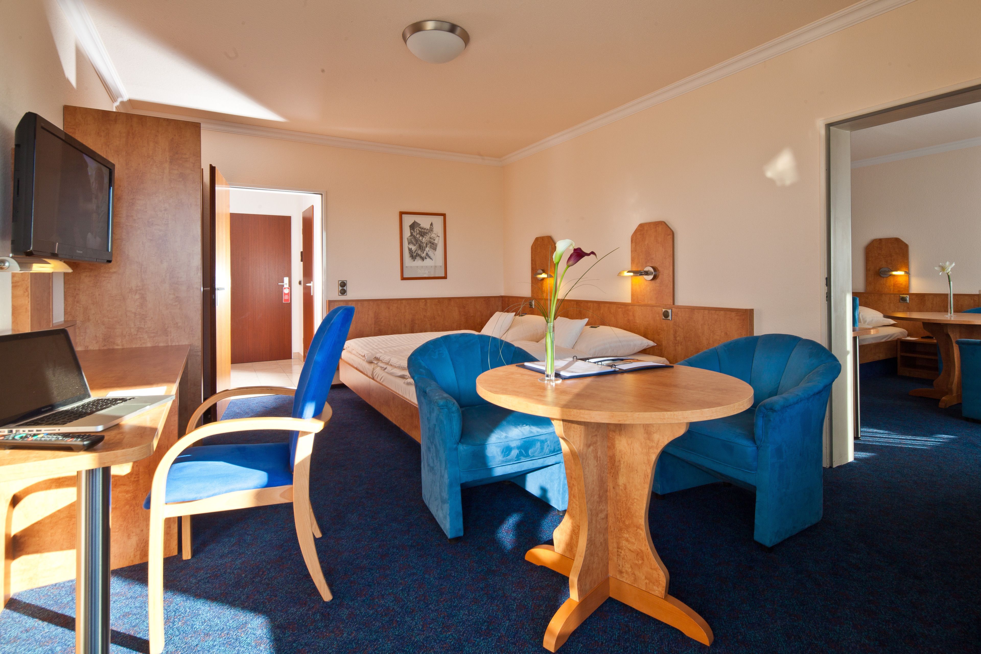 A hotel room with a bed, two blue armchairs, a wooden table, a desk with a laptop, a TV, and a framed picture on the wall.