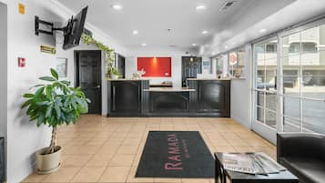A reception area with a black reception desk, a large potted plant, a red wall, and a black rug with 'RAMADA' written on it.