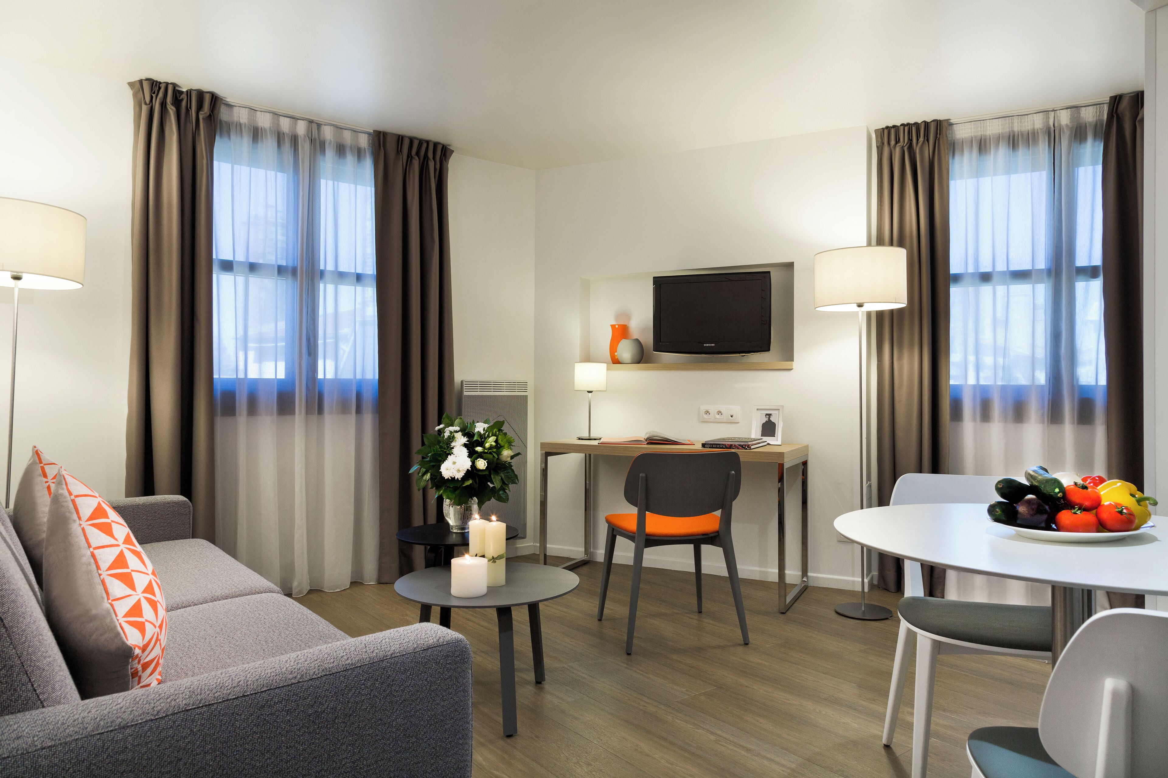 A modern hotel room with a sofa, a dining table, a TV, and a small desk.
