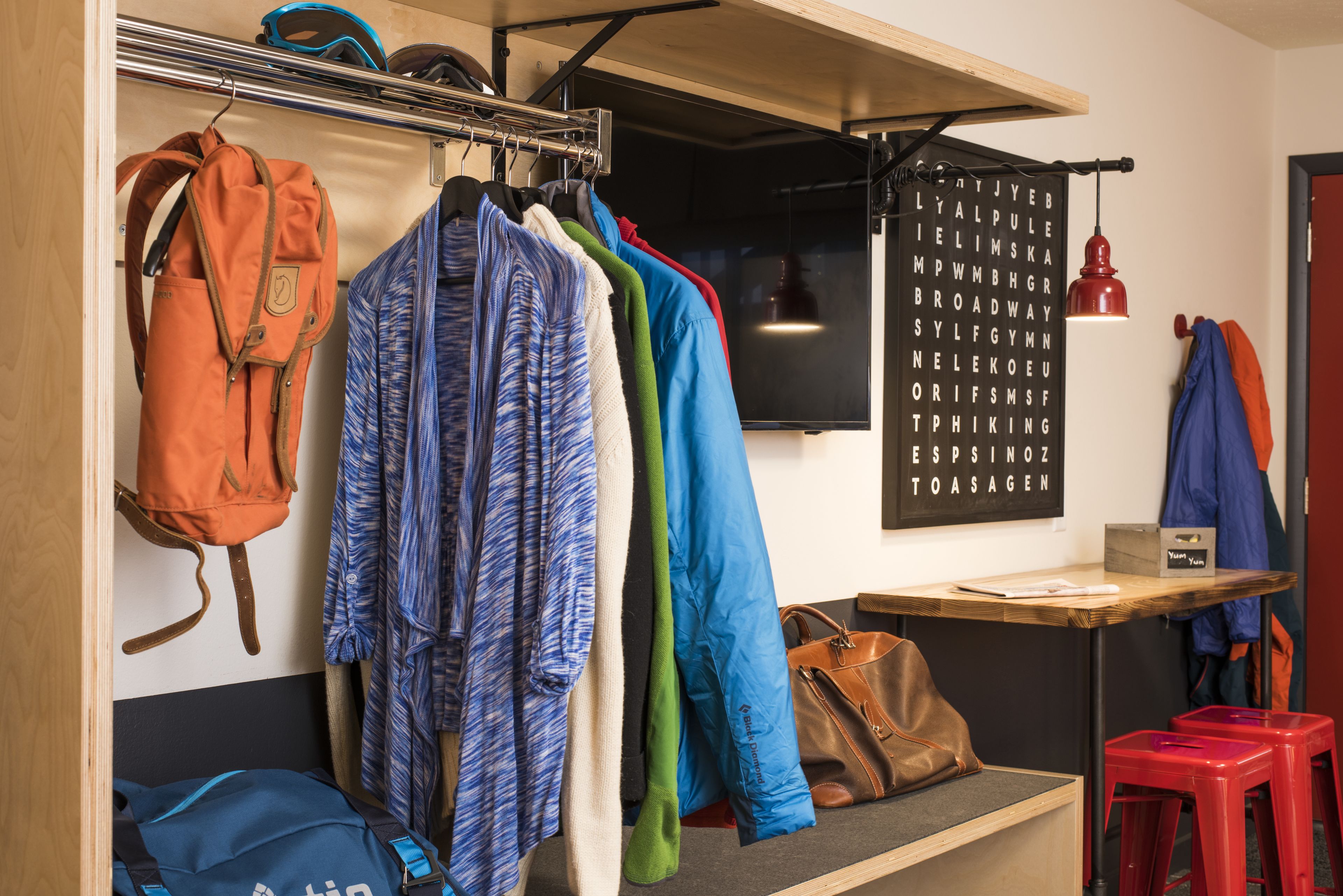 A room with a wooden bench, a hanging coat rack with various jackets, a backpack, and a red stool.