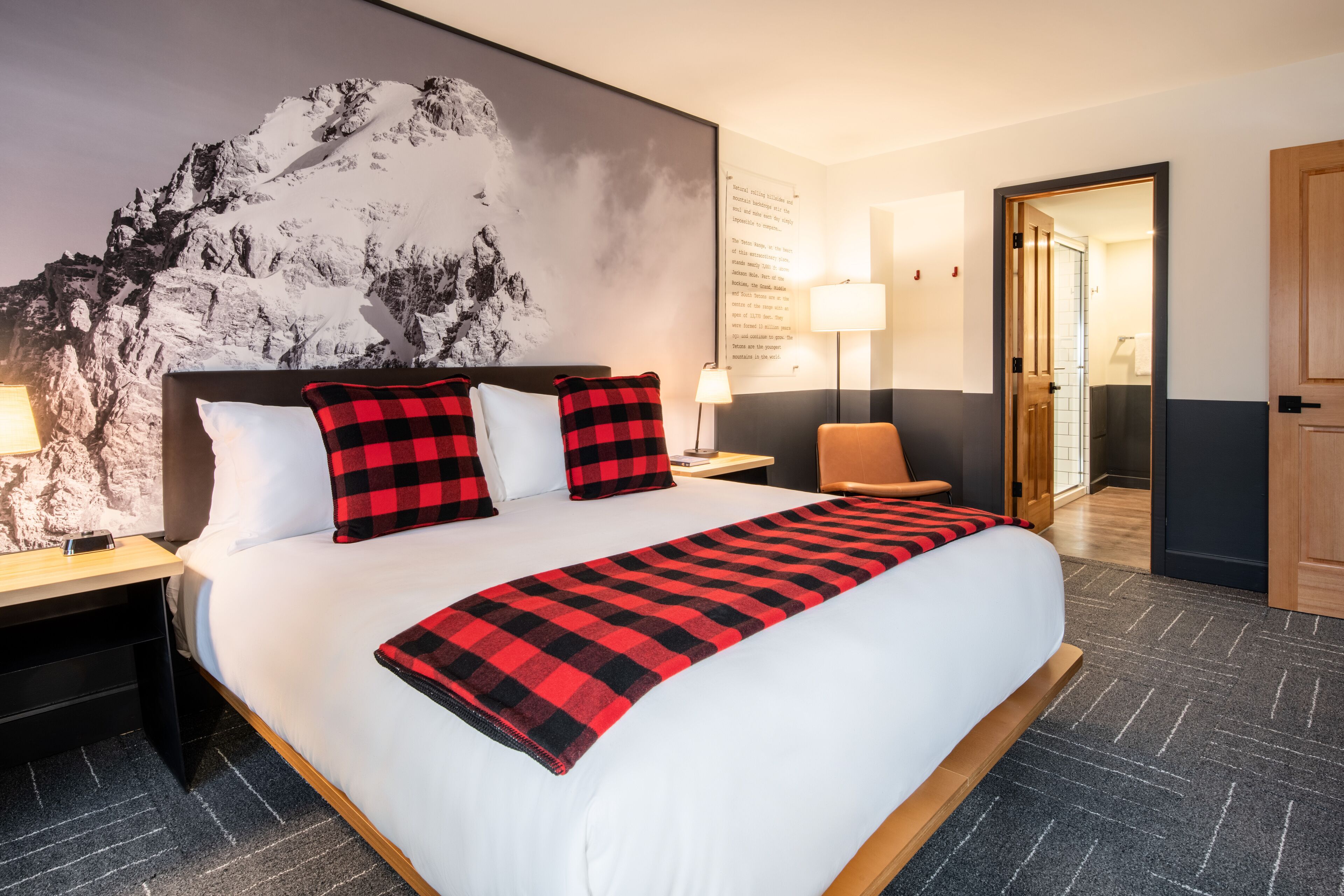 A hotel room with a large bed, two nightstands, a chair, a wall-mounted lamp, and a door leading to another room.