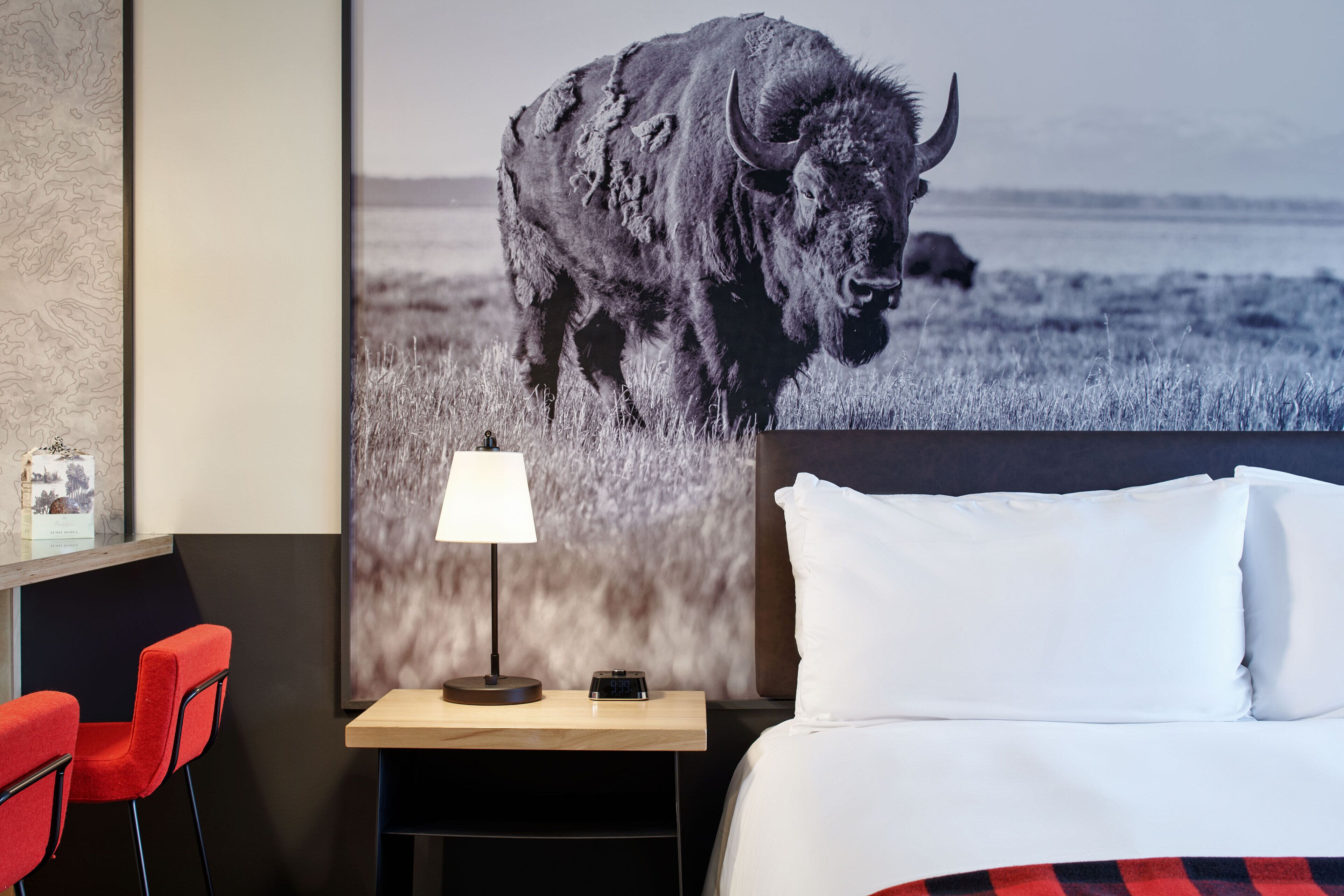 Quarto de hotel com uma cama, uma mesa de cabeceira com abajur, uma cadeira vermelha e um grande cartaz na parede de um bisão.