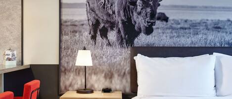 Quarto de hotel com uma cama, uma mesa de cabeceira com abajur, uma cadeira vermelha e um grande cartaz na parede de um bisão.