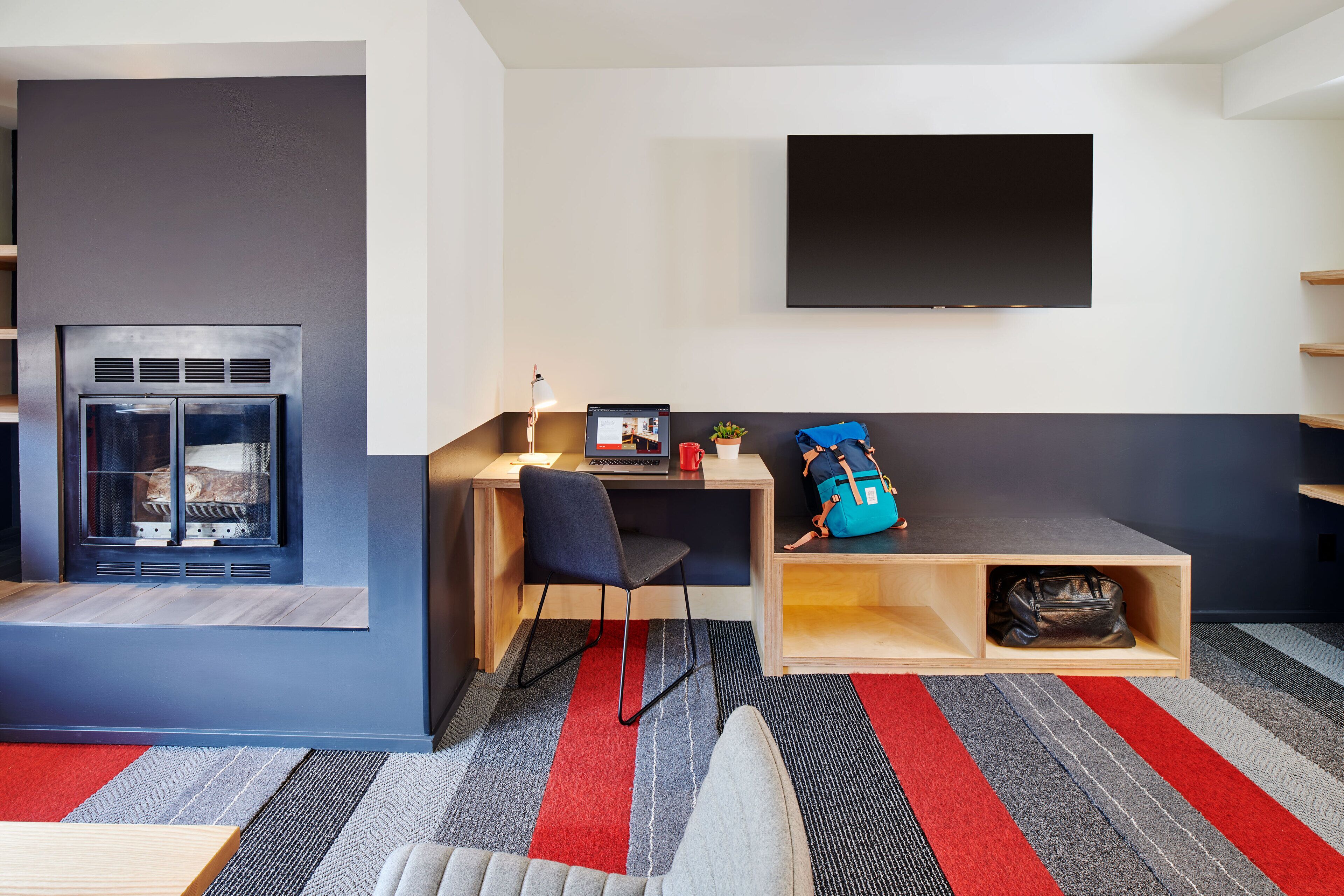 Ett modernt rum med en öppen spis, ett skrivbord med en dator, en tv som är monterad på väggen och en röd och grå randig matta.