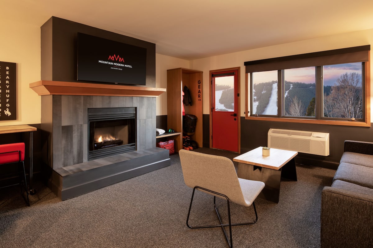 A modern living room with a fireplace, a television mounted above it, a red door, and a large window with a view of a snowy landscape.