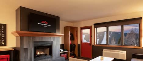 A modern living room with a fireplace, a television mounted above it, a red door, and a large window with a view of a snowy landscape.