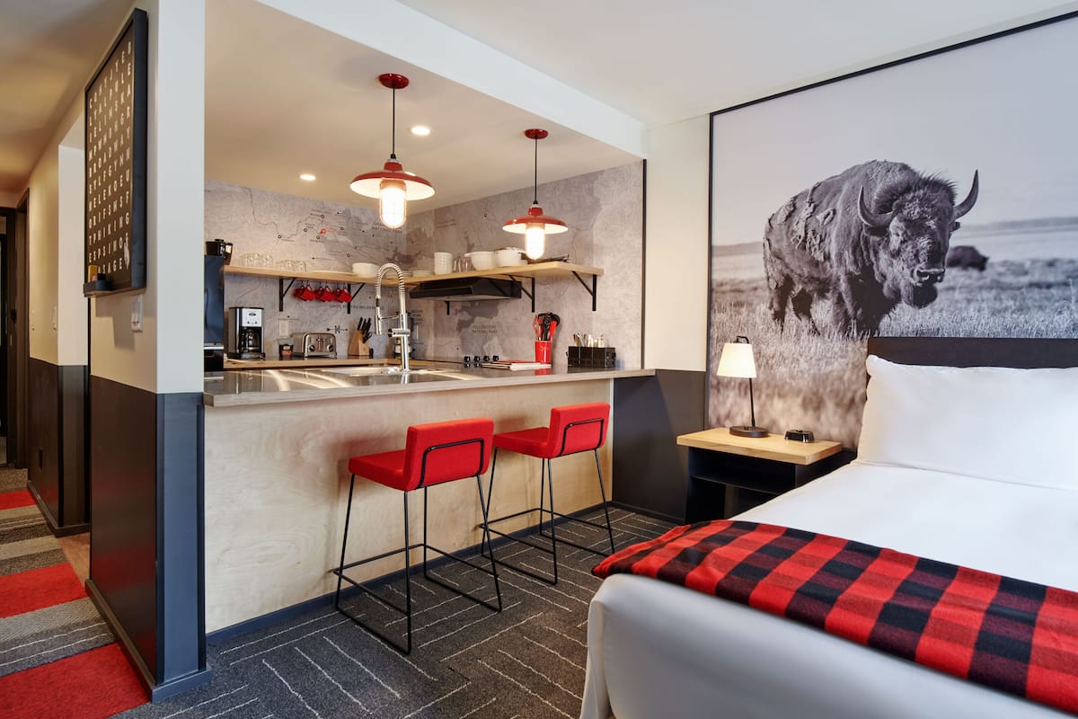 A modern hotel room with a large bed, a red and black checkered throw, a map on the wall, a kitchen area with a bar, and a bull painting.
