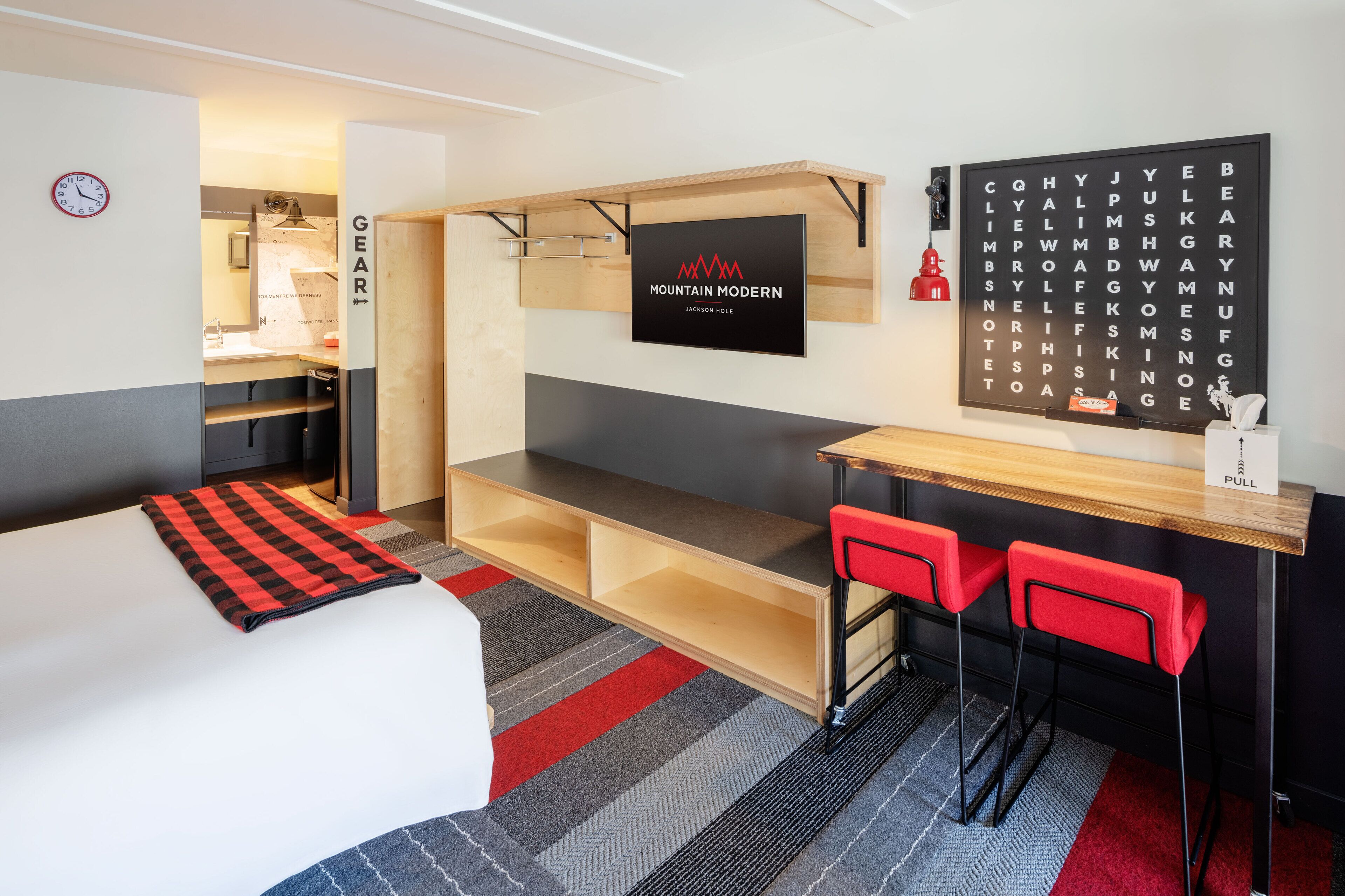 Een moderne hotelkamer met een bed, een bureau met twee rode stoelen, een wandtelevisie en een kruiswoordraadsel aan de muur.