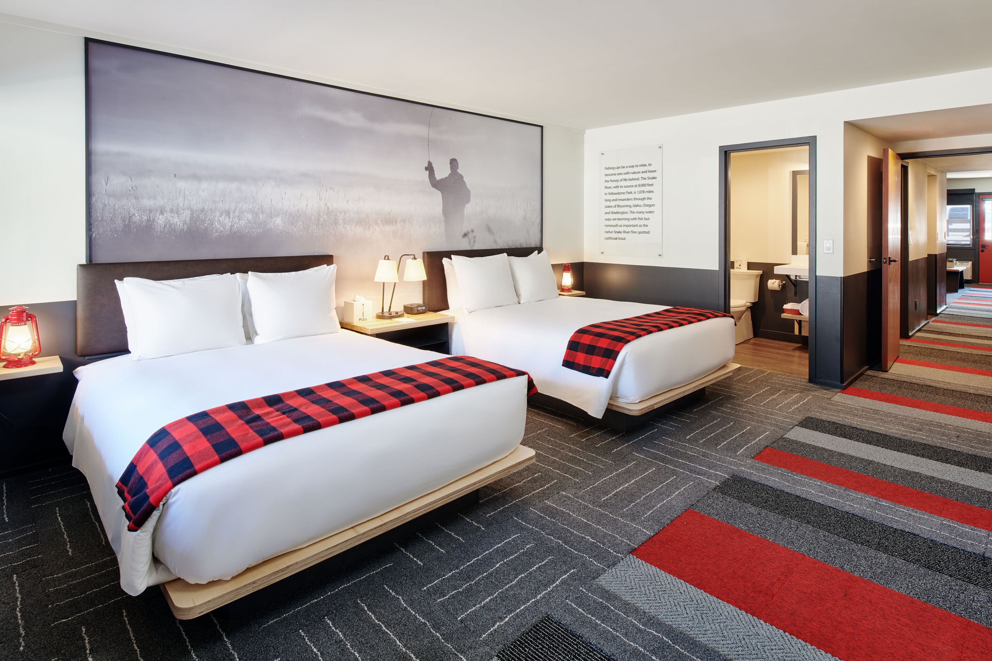 Une chambre d’hôtel avec deux lits, une grande œuvre d’art au mur, une couverture à carreaux rouges et noirs sur l’un des lits, et un sol recouvert de moquette.
