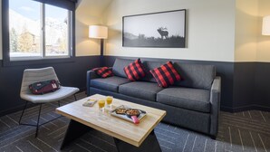 A modern room with a grey sofa, red and black pillows, a wooden coffee table with breakfast items, a chair with a bag, and a mountain view through a large window. - Mountain Modern Jackson Hole (Jackson)