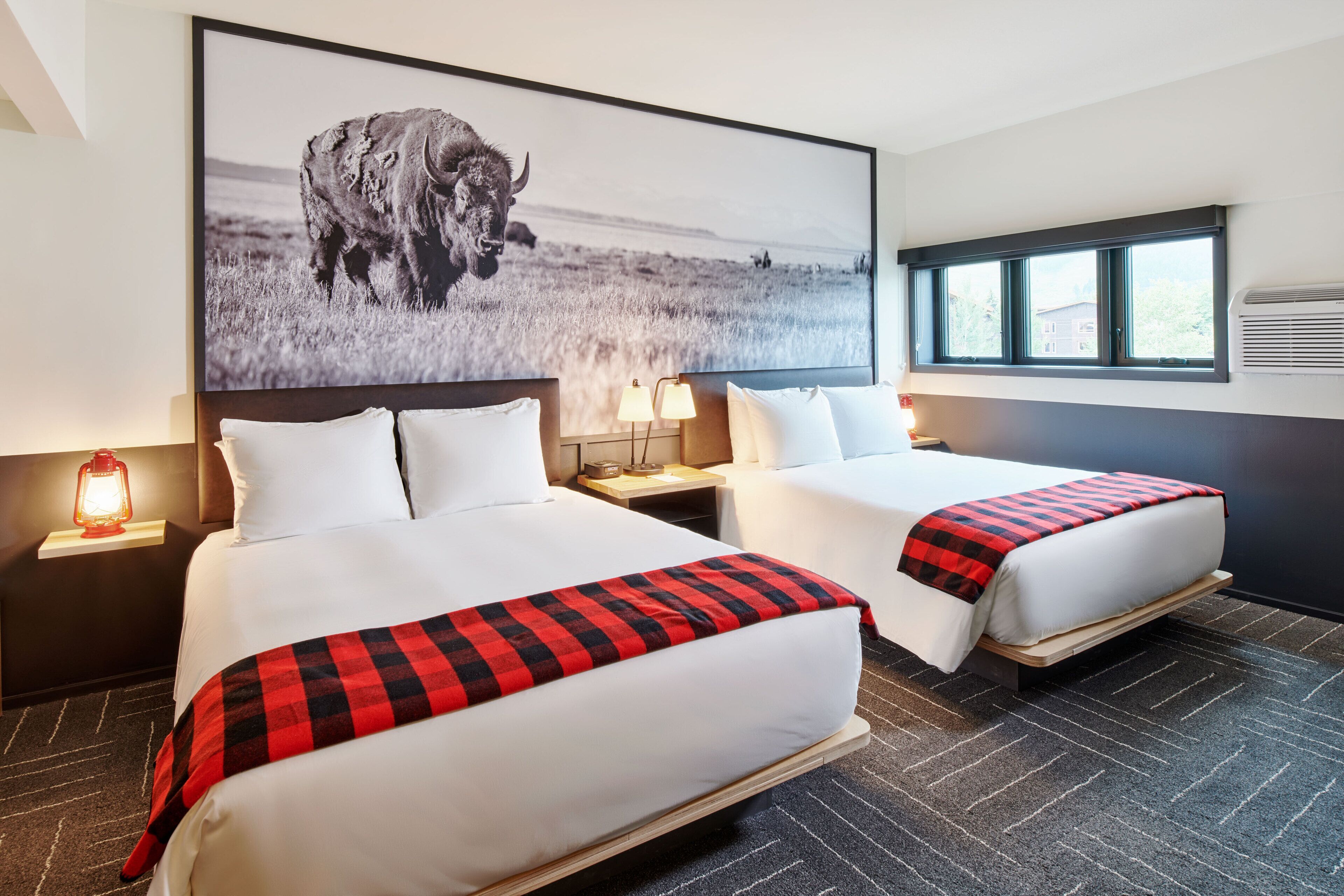 A hotel room with two beds, a large wall-mounted picture of a bison, a window with blinds, and a nightstand with a lamp.