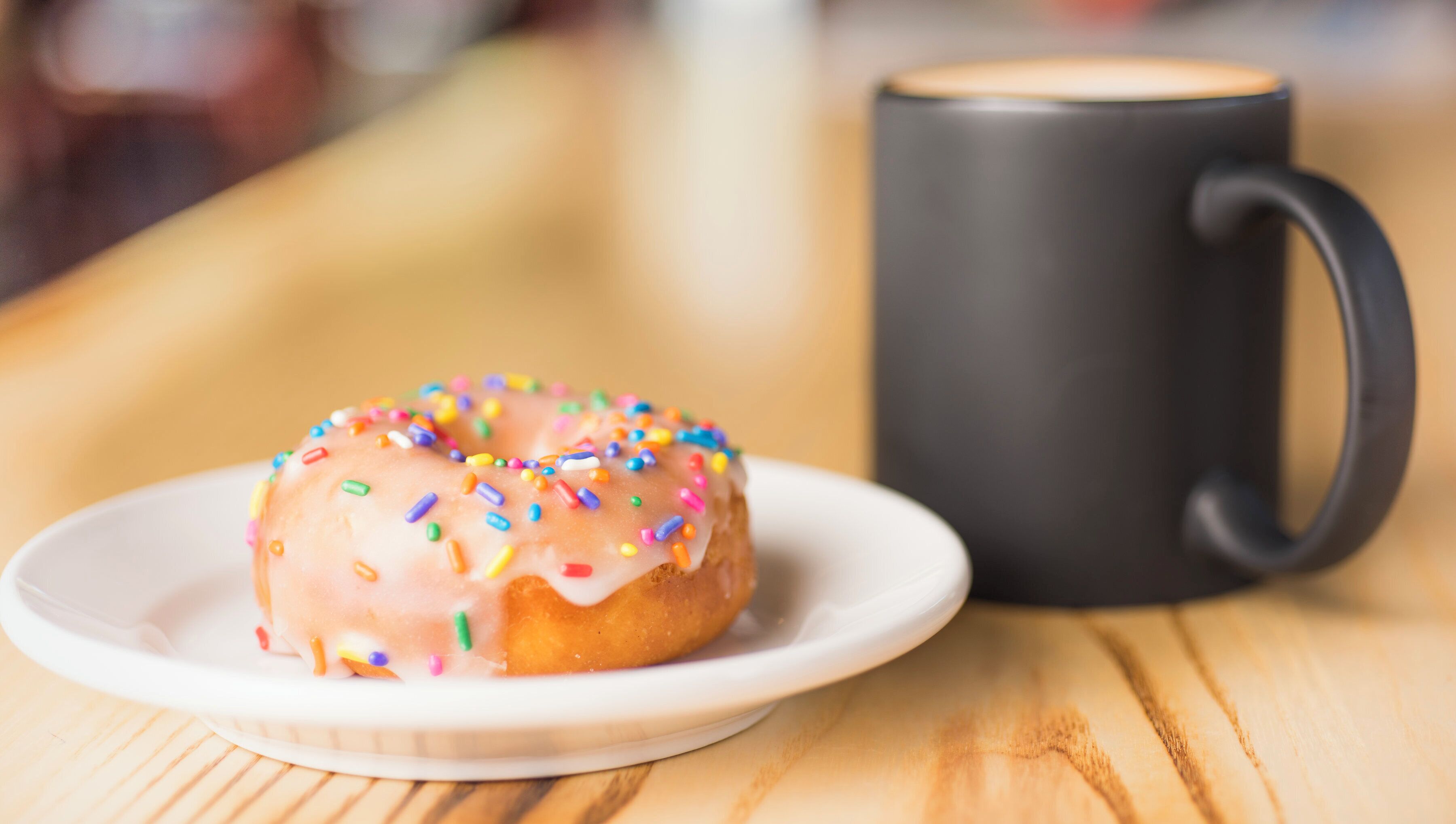 Un donut con codette colorate su un piatto bianco, accanto a una tazza nera su un tavolo di legno.