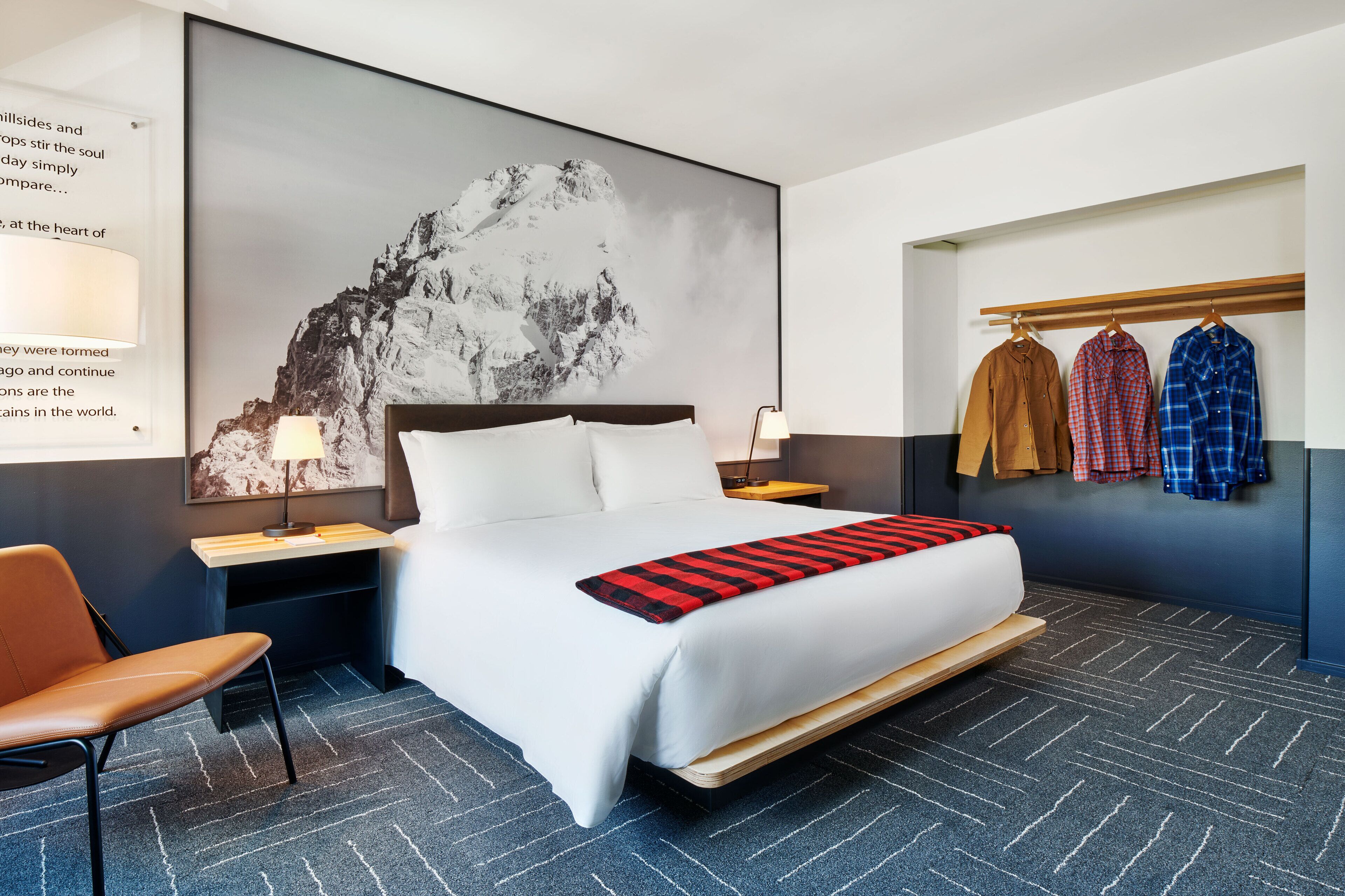 A hotel room with a large bed, a desk, a chair, and a wall-mounted coat rack with three jackets.