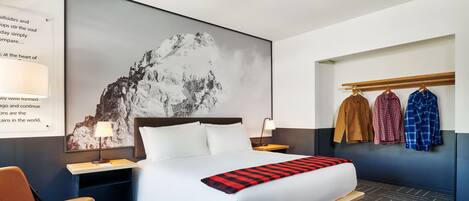 A hotel room with a large bed, a desk, a chair, and a wall-mounted coat rack with three jackets.