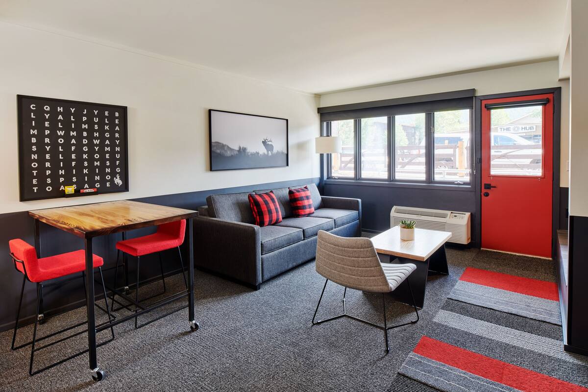 A modern room with a sofa, two red chairs, a wooden table, a lamp, and a window with a view of a building.