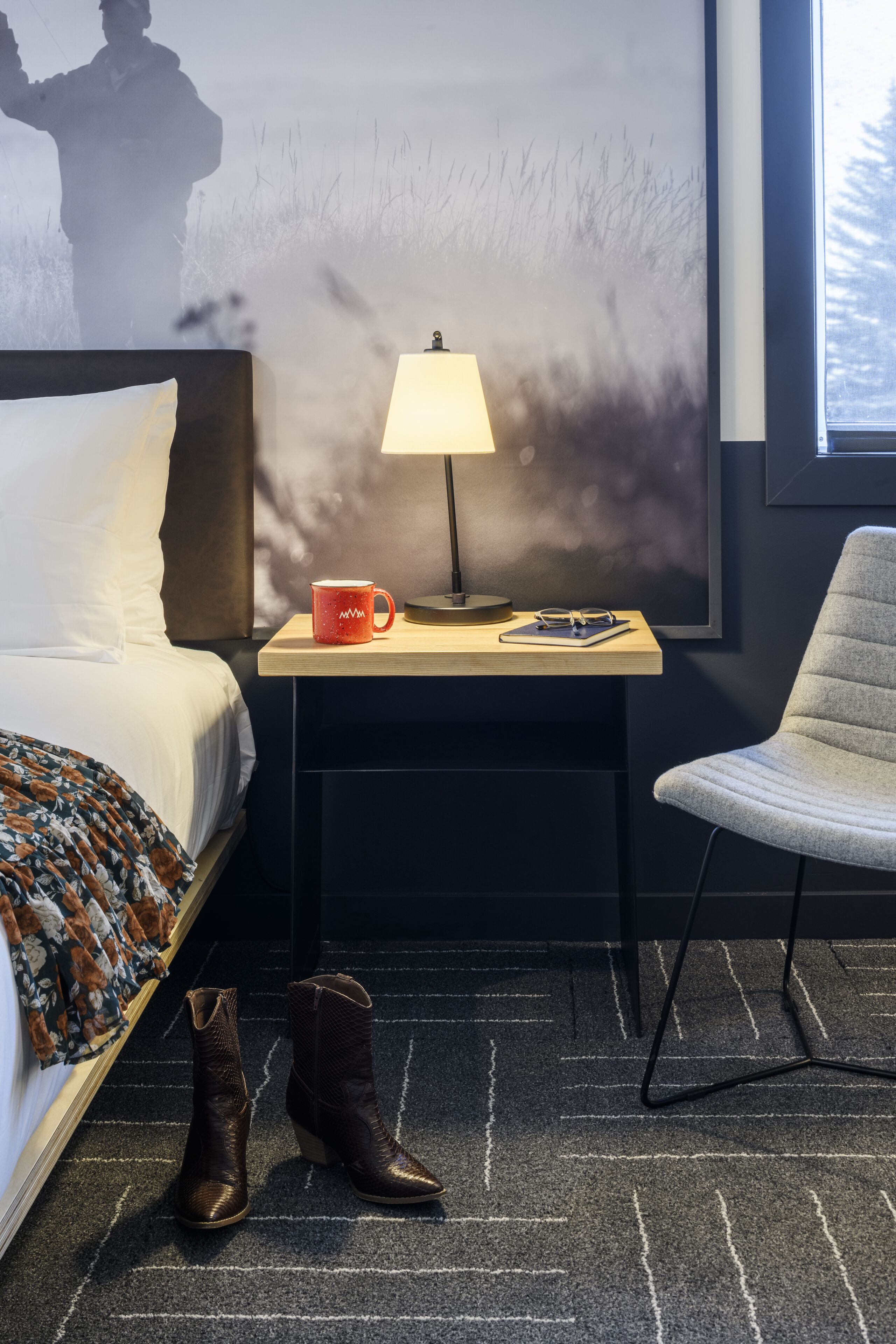 A hotel room with a bed, a nightstand with a lamp, a chair, and a pair of boots on the floor.