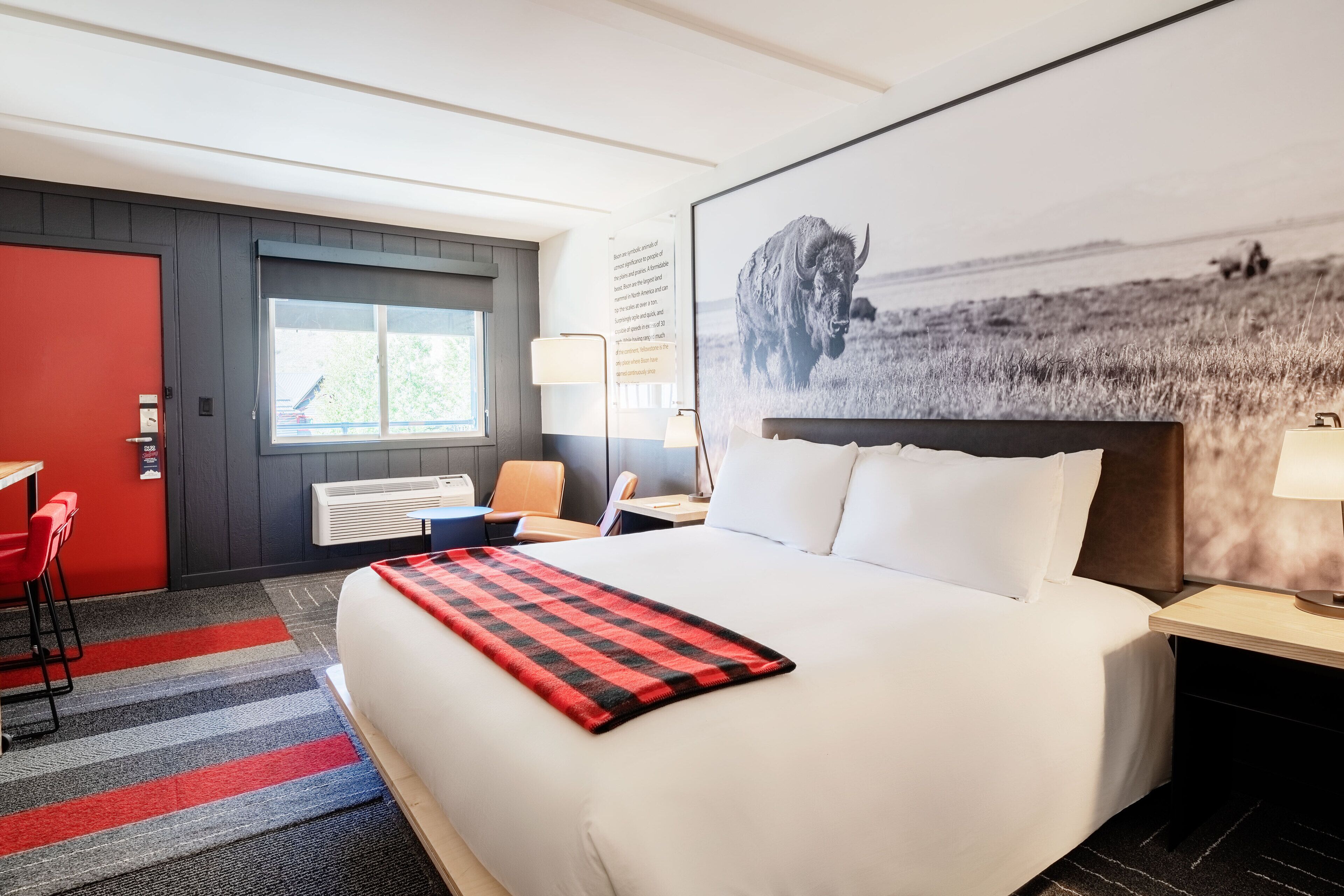 A hotel room with a large bed, a red and black checkered throw, a red door, a window with a view of a field, and a mounted picture of a bison.