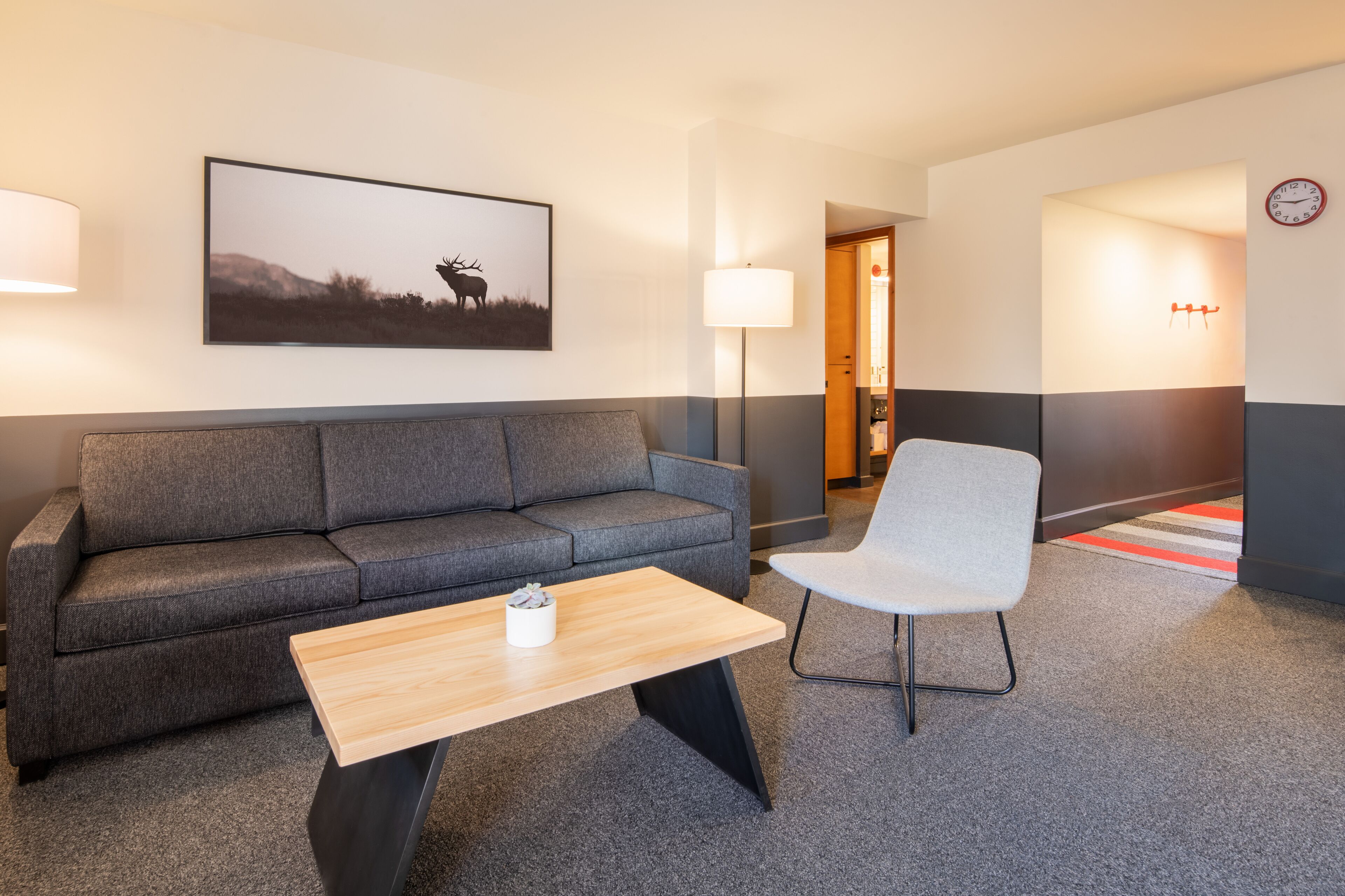A modern hotel room with a grey sofa, a wooden coffee table, a white armchair, a framed picture of a deer, a floor lamp, and a clock on the wall.