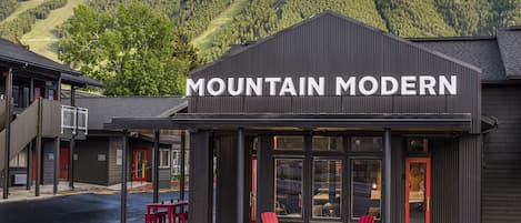 A building named "Mountain Modern" with a modern architectural design, featuring a prominent sign, outdoor seating area with red chairs and tables, and a view of a forested mountain in the background.