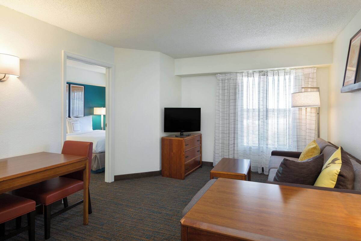 Een hotelkamer met een houten eettafel, een bank met kussens, een televisie op een houten dressoir en een raam met gordijnen.
