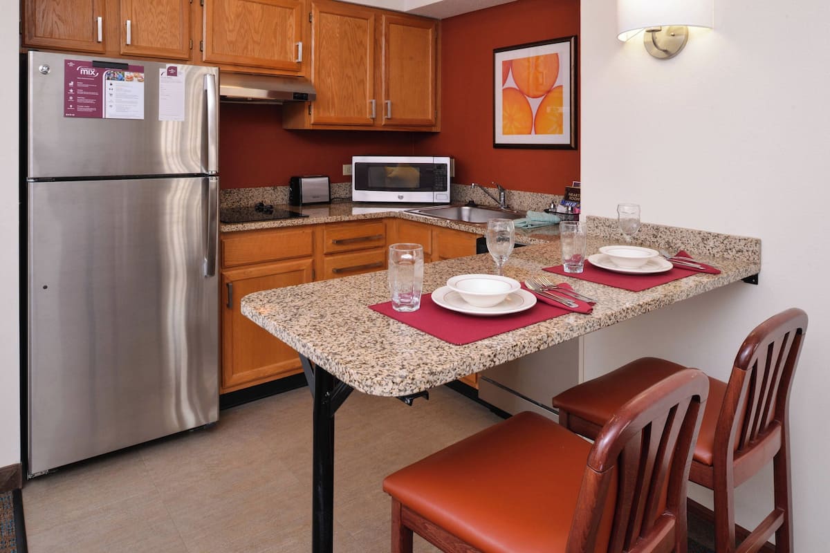 A compact kitchen with a stainless steel refrigerator, microwave, and granite countertop. There are wooden cabinets and a dining table with chairs.
