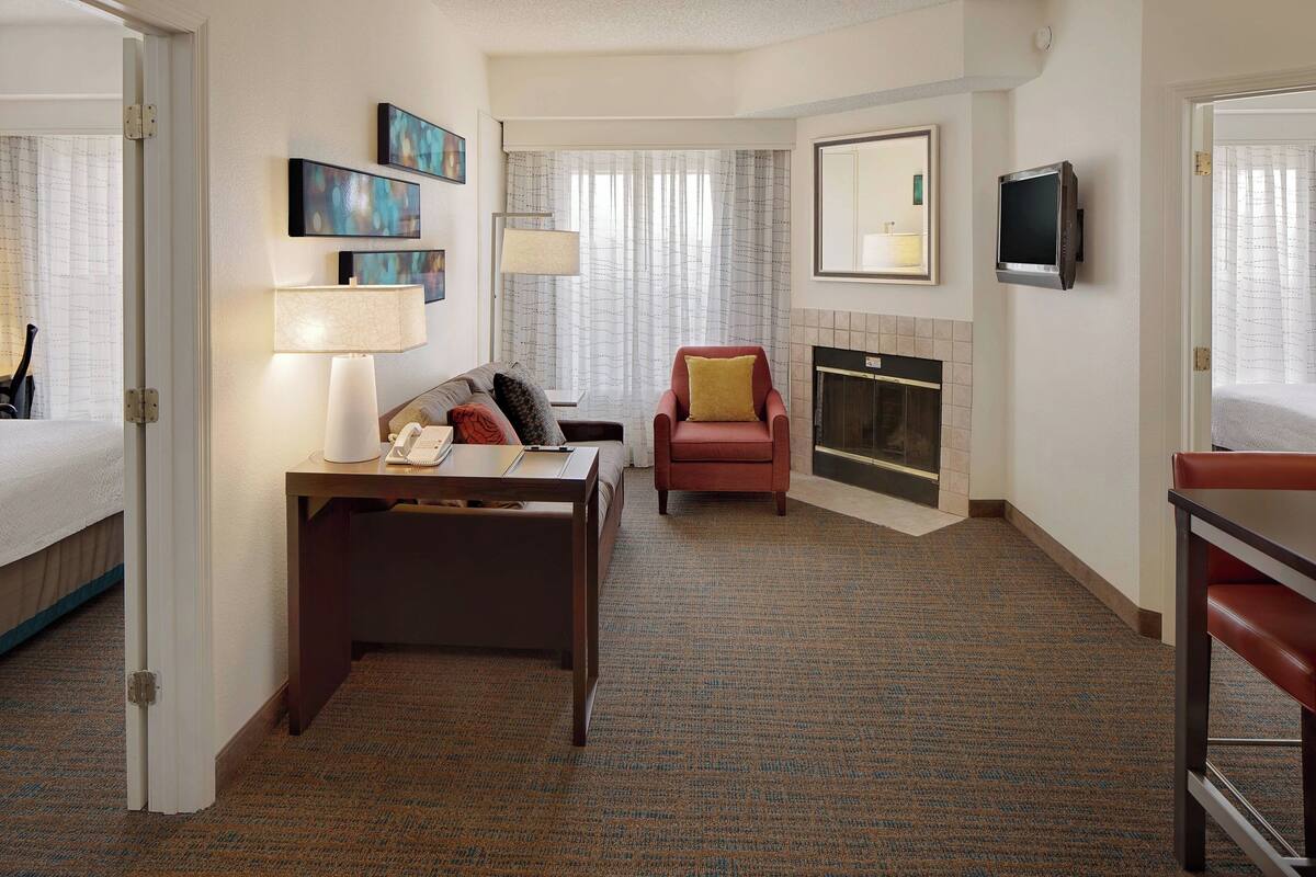 A hotel room with a bed, a desk with a telephone, a red armchair, a fireplace, and a television mounted on the wall.