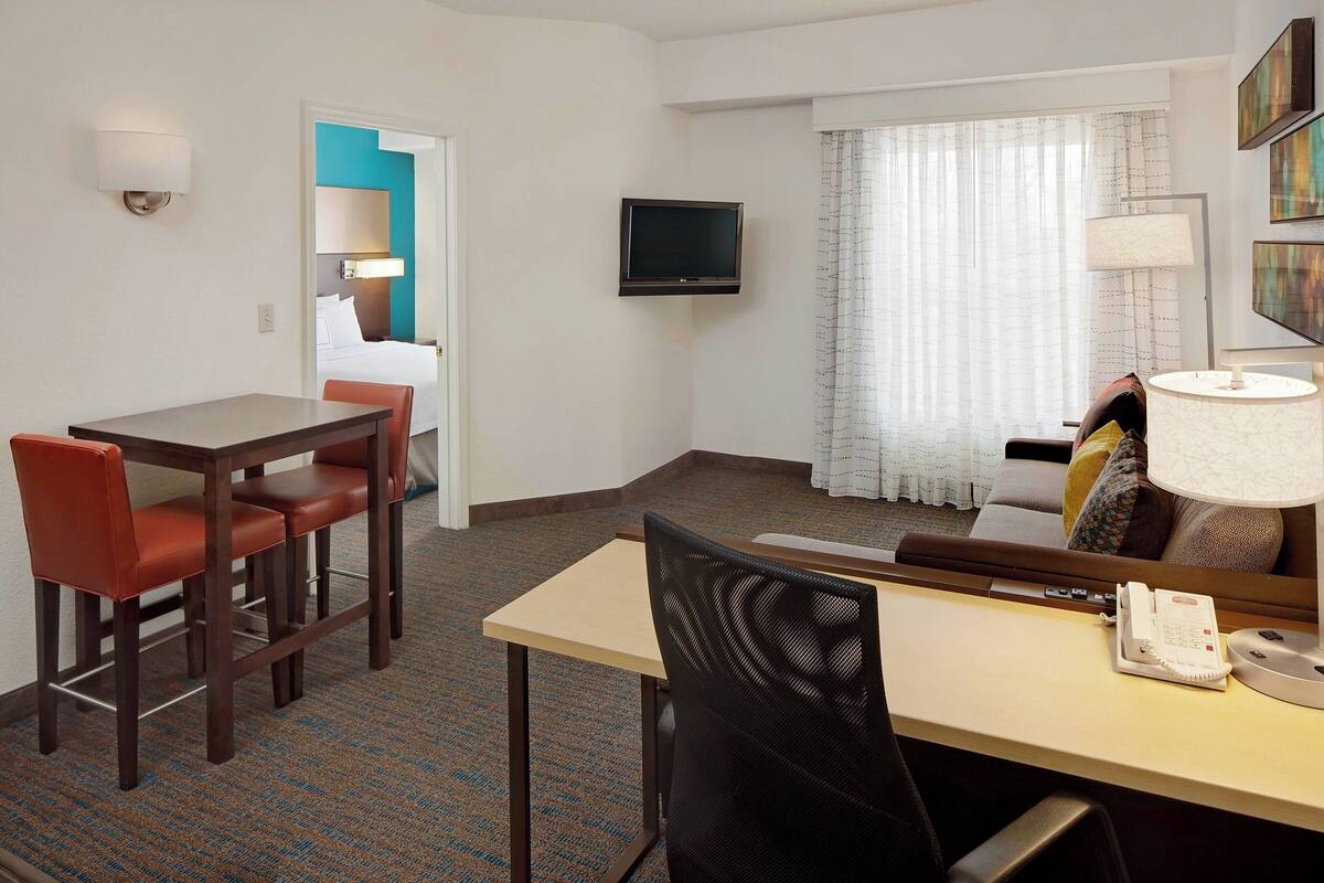 A hotel room with a desk, two chairs, a television, a bed, a window with curtains, and a lamp.