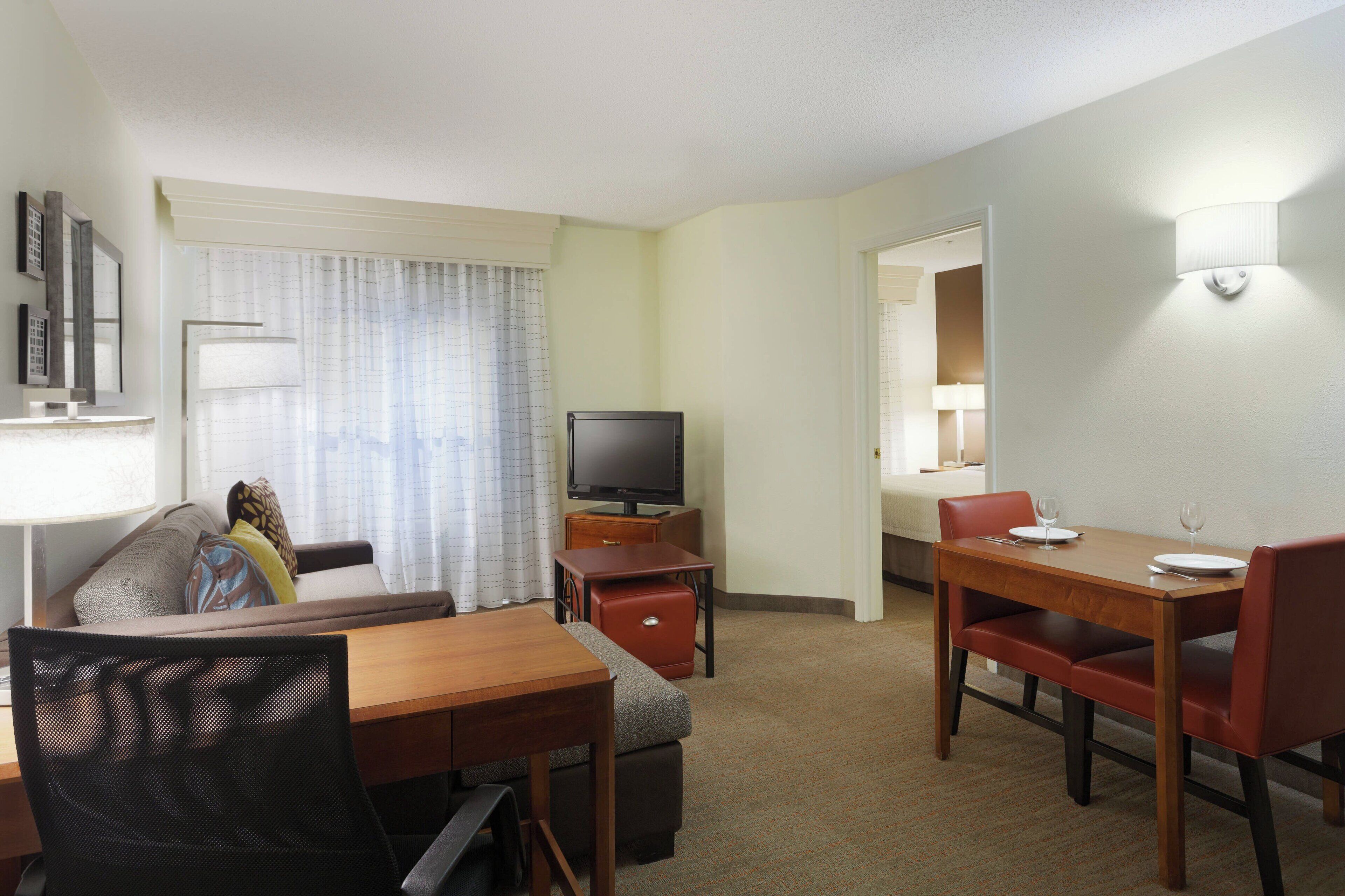 A hotel room with a wooden dining table, red chairs, a television, a sofa, and a window with curtains.