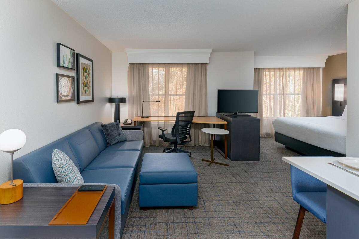 Quarto de hotel com um sofá azul, uma escrivaninha com cadeira, uma cama e uma televisão.