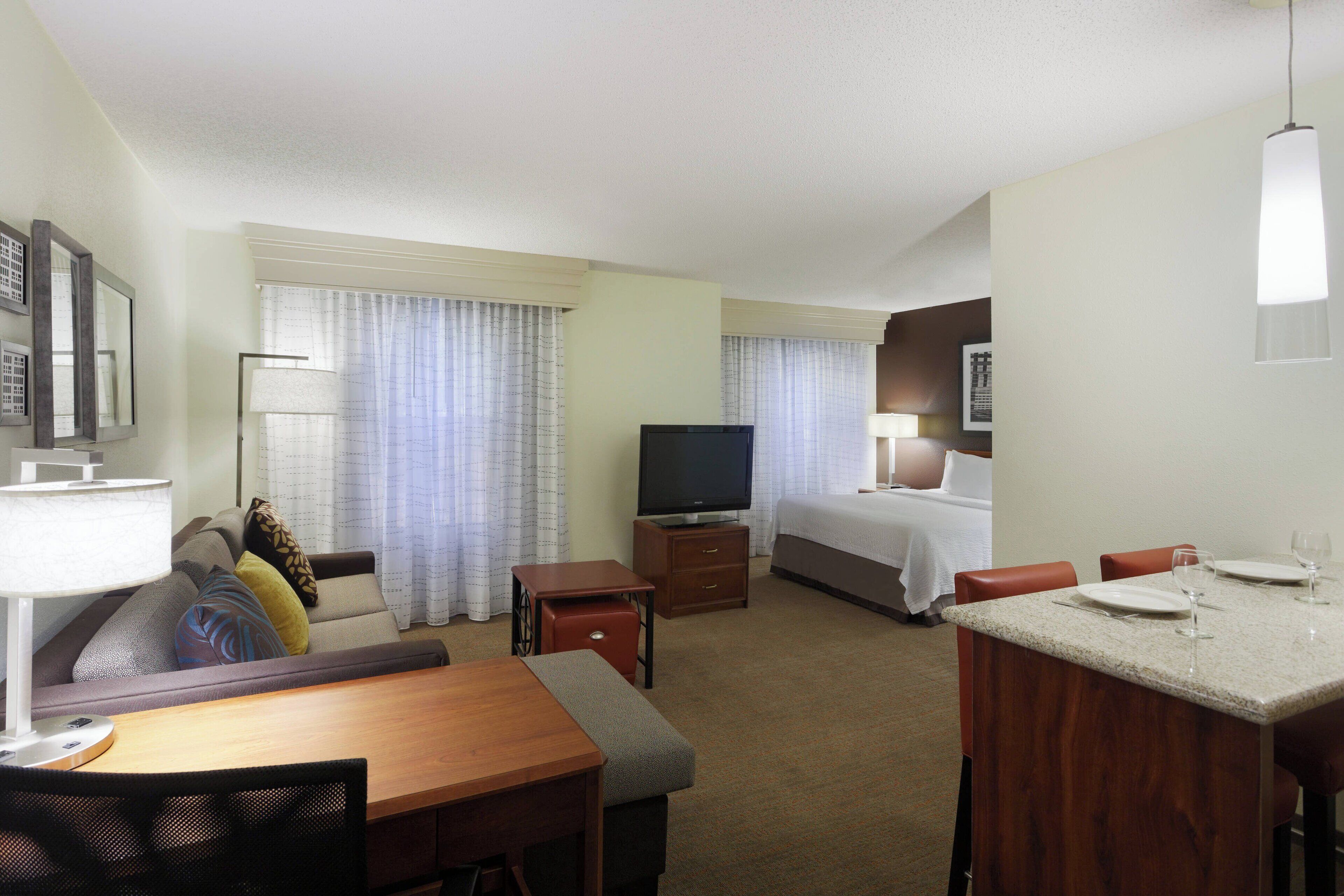 A hotel room with a bed, a TV, a desk with a chair, a dining table with chairs, and a window with curtains.