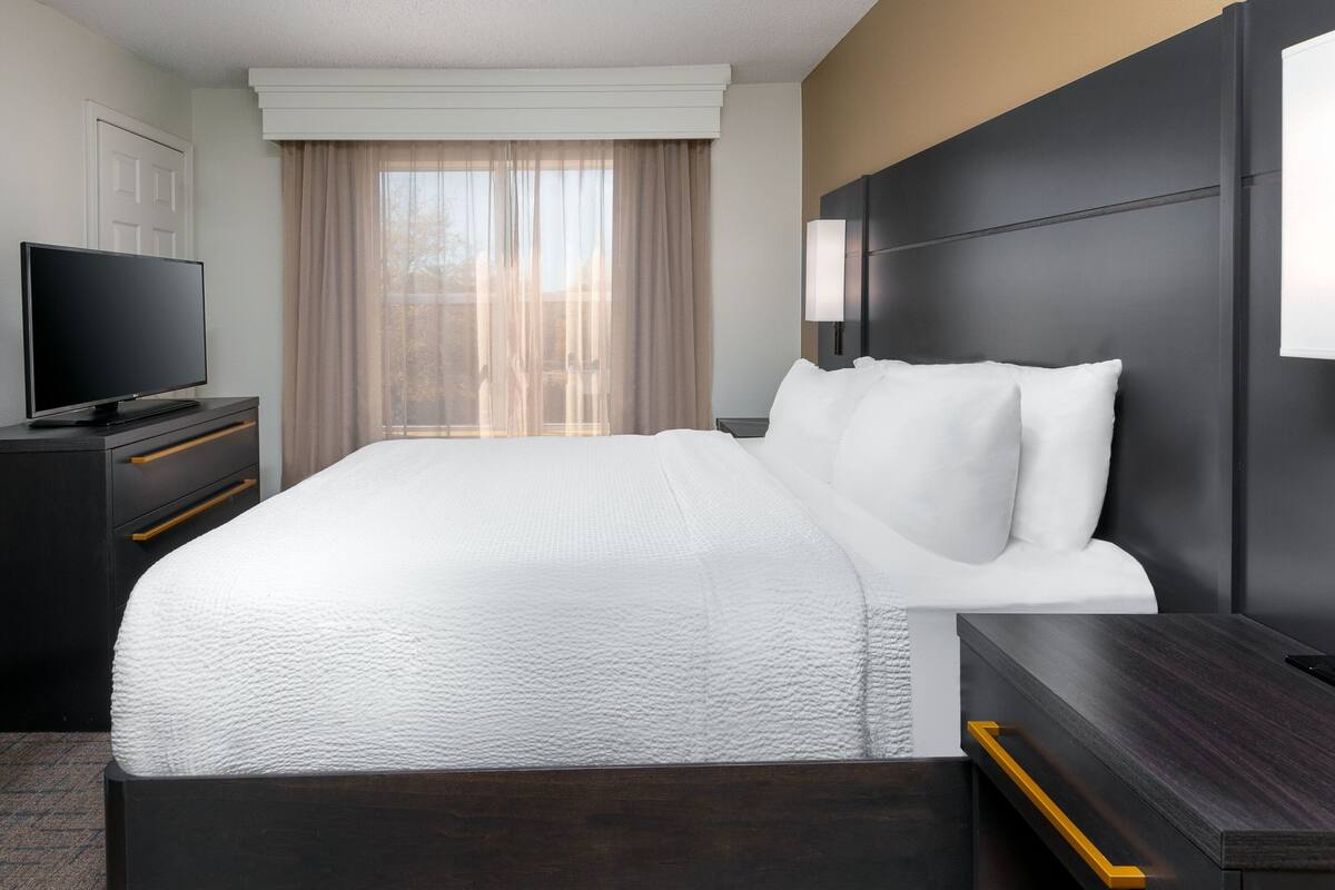 Quarto de hotel com uma cama grande, televisão em uma cômoda, mesa de cabeceira e janela com cortinas.