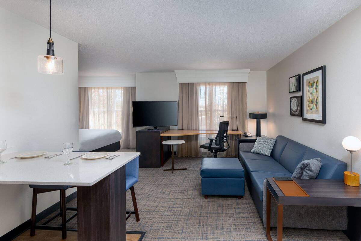 Quarto de hotel moderno com televisão de tela plana, mesa de jantar com cadeiras, sofá, escrivaninha e luminária.