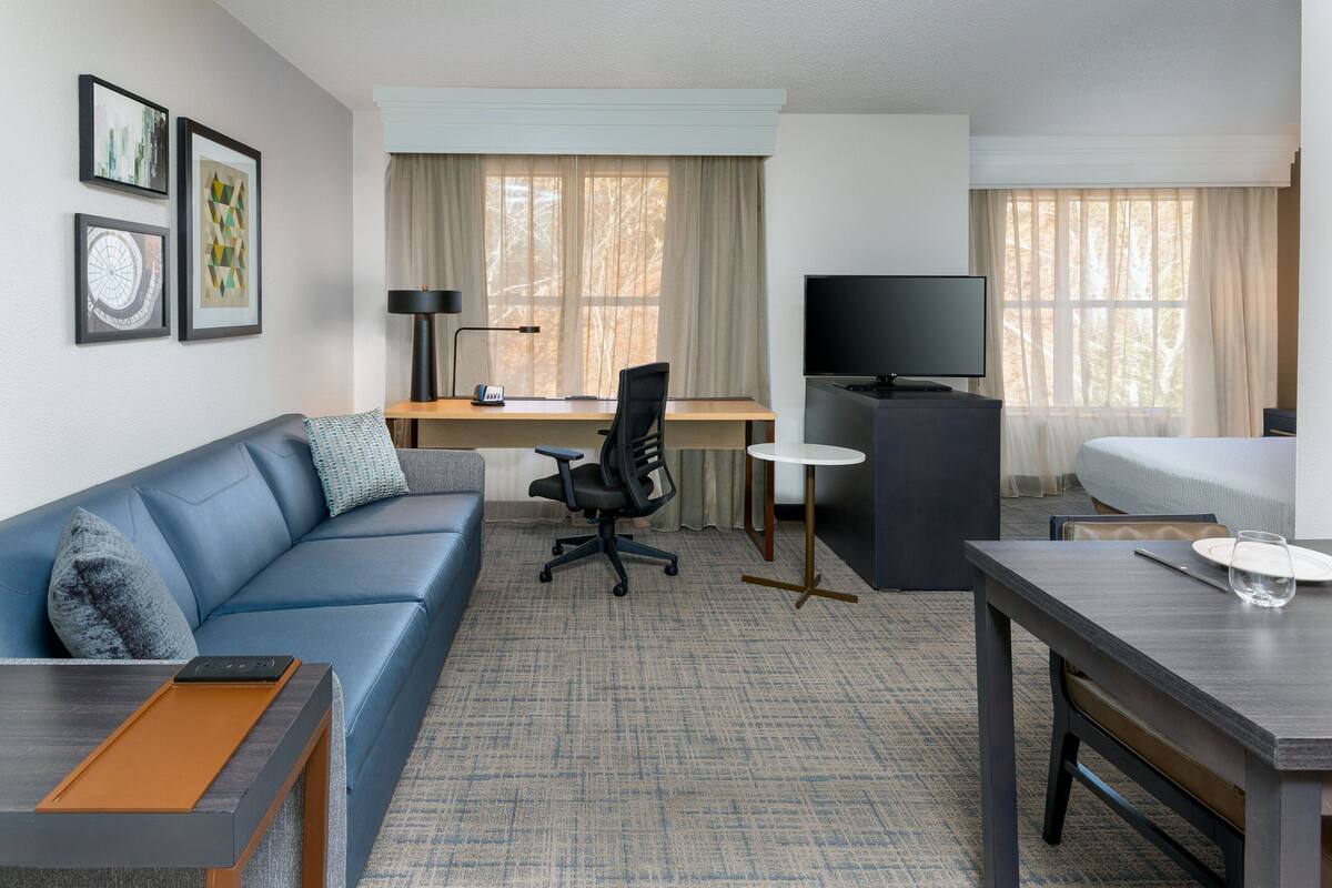Quarto de hotel com um sofá azul, uma escrivaninha com computador, televisão e cama.