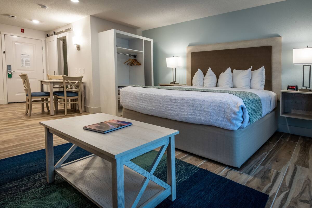 A hotel room with a bed, bedside table, and a desk with chairs. There is a closet with hanging clothes and a window with curtains.
