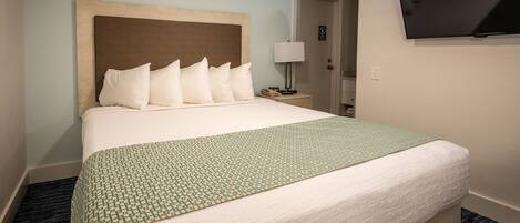 A hotel room with a large bed, a flat-screen TV mounted on the wall, and a bedside table with a lamp.