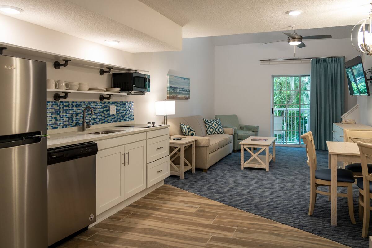A modern kitchen with a dishwasher, microwave, and stainless steel refrigerator. There is a dining area with a table and chairs, and a sofa with patterned cushions. A ceiling fan and a window with curtains are visible.