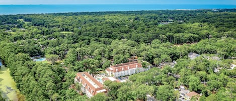 An aerial view of a large estate with a central building surrounded by dense forest, a swimming pool, and a view of the ocean.