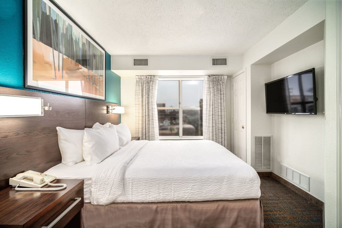 A hotel room with a large bed, a bedside telephone, a television mounted on the wall, and a window with curtains.