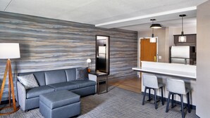 A modern hotel room with a grey sectional sofa, a small dining area with two chairs, a bar counter, a large mirror, and a floor lamp. - Residence Inn by Marriott Rochester West/Greece (Rochester)