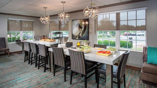 A dining area with a long table, chairs, and a view of the parking lot through large windows. - Residence Inn By Marriott Louisville Northeast (Louisville)