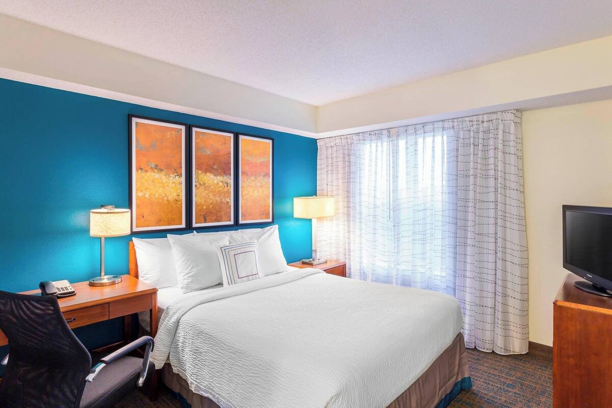 A hotel room with a bed, two bedside tables, a desk with a chair, a television, and a window with curtains.