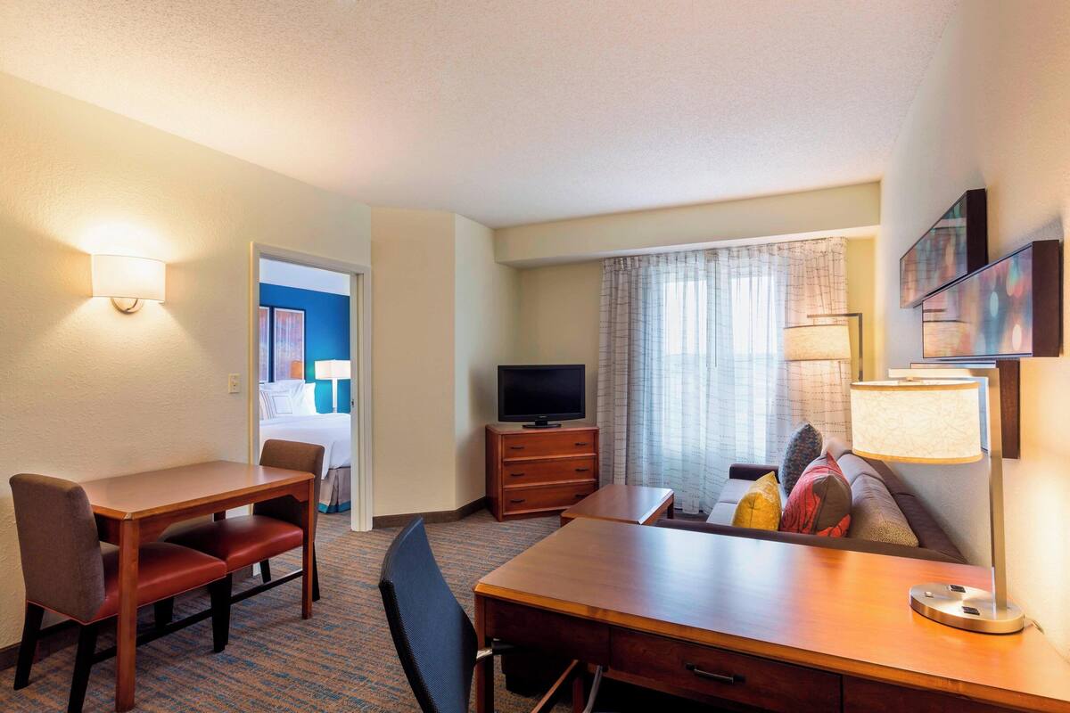 A hotel room with a wooden desk, two chairs, a television, a sofa, and a lamp.