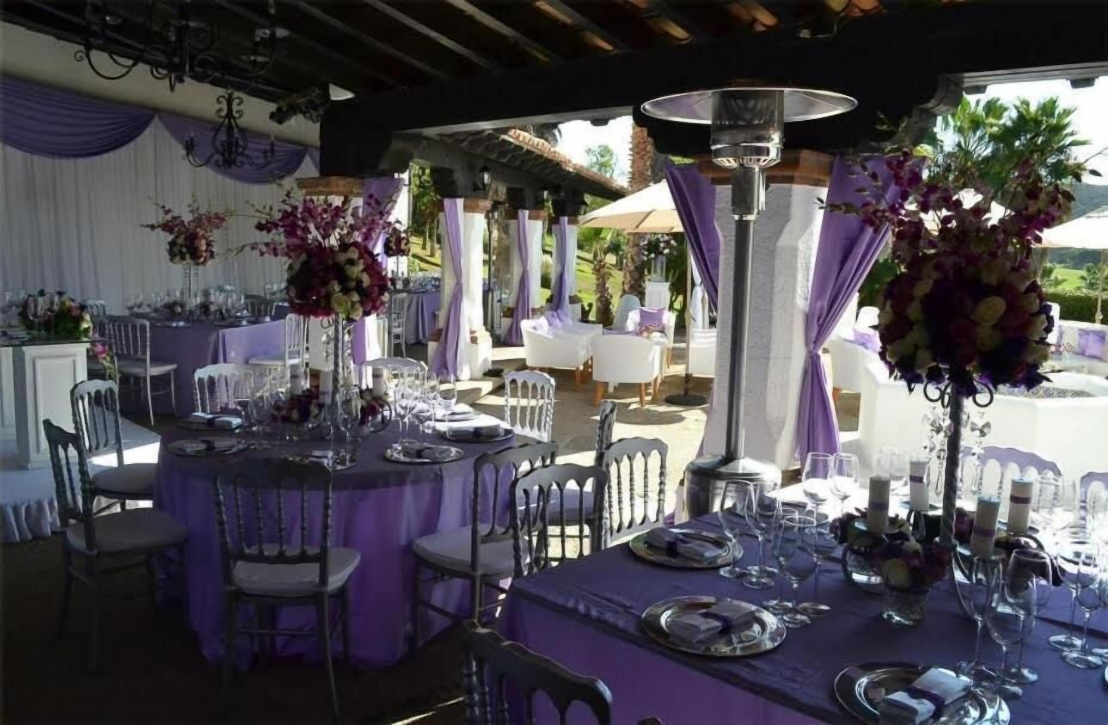 Un espace de restauration en plein air, avec des tables dressées pour un événement formel, agrémenté de nappes violettes et de centres de table floraux.