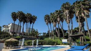 A pool area with blue lounge chairs, a pool, palm trees, and a building in the background. - Real del Mar Hotel & Golf Resort (Tijuana)